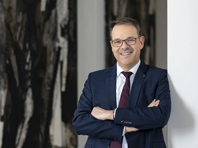 Generalintendant und Geschäftsführer der Städtische Theater Chemnitz gGmbh in blauem Anzug und gestreifter Krawatte mit verschränkten Armen lehnt an einer weißen Wand vor abstrakten Gemälden.